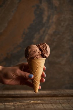 cropped view of woman holding chocolate ice cream cone over wooden table clipart