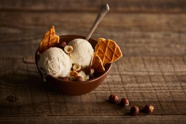 delicious ice cream with pieces of waffle, spoon and hazelnuts in bowl on wooden surface clipart