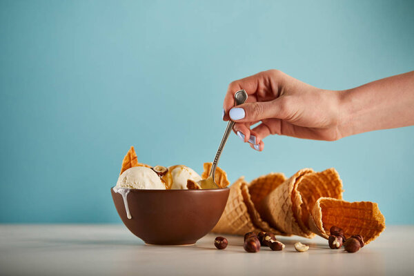 partial view of woman with spoon, bowl of ice cream, hazelnuts and waffle cones on blue