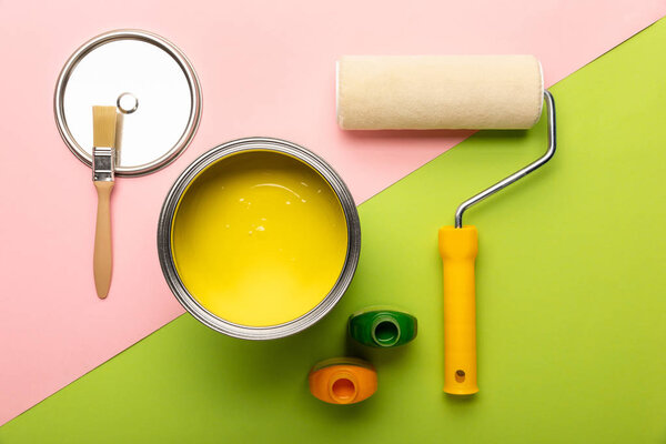 top view of tin with yellow paint, bottles, brush and paint roller on pink and green surface