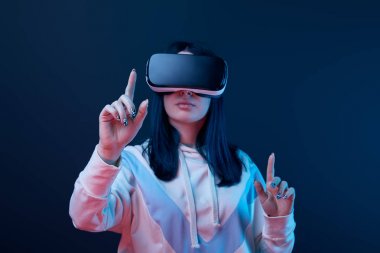 selective focus of young brunette woman wearing virtual reality headset and pointing with fingers on blue  clipart