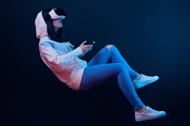 KYIV, UKRAINE - APRIL 5, 2019: Brunette woman levitating and holding joystick while playing video game and wearing virtual reality headset on blue   clipart
