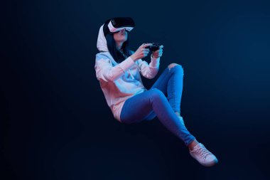 KYIV, UKRAINE - APRIL 5, 2019: Young woman in virtual reality headset levitating and holding joystick on blue   clipart