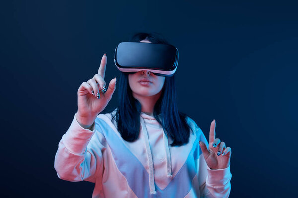 selective focus of young brunette woman wearing virtual reality headset and pointing with fingers on blue 