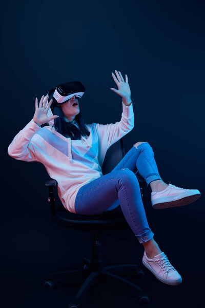 shocked young woman gesturing and using virtual reality headset while sitting on chair on blue 