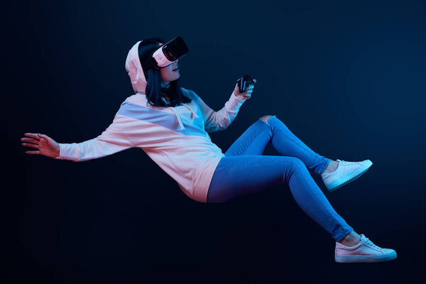 KYIV, UKRAINE - APRIL 5, 2019: Surprised woman in virtual reality headset levitating and holding joystick on blue  