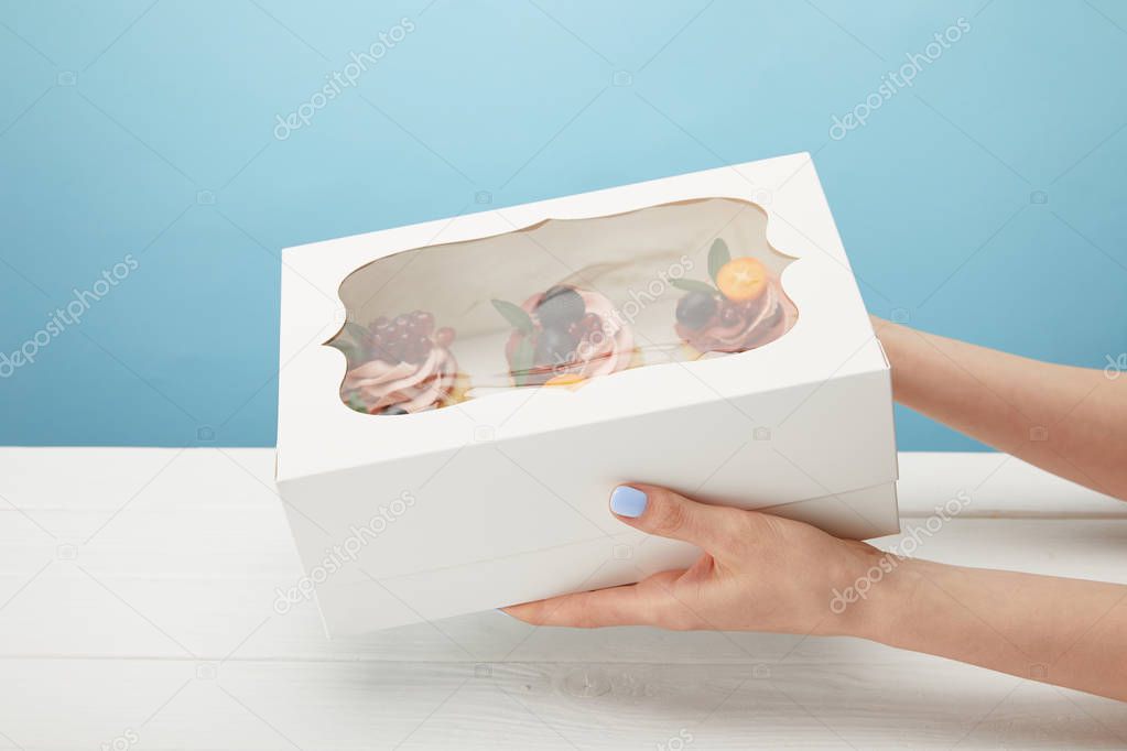 partial view of woman holding box with cupcakes isolated on blue