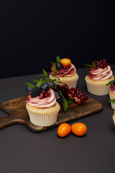 sweet cupcakes with fruits and berries and cutting board on grey surface isolated on black