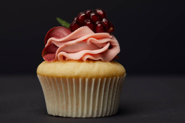 sweet cupcake with cream and garnet on grey surface isolated on black