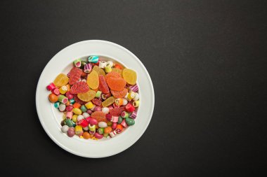 top view of delicious multicolored caramel candies and citrus jellies in white plate on black background with copy space clipart