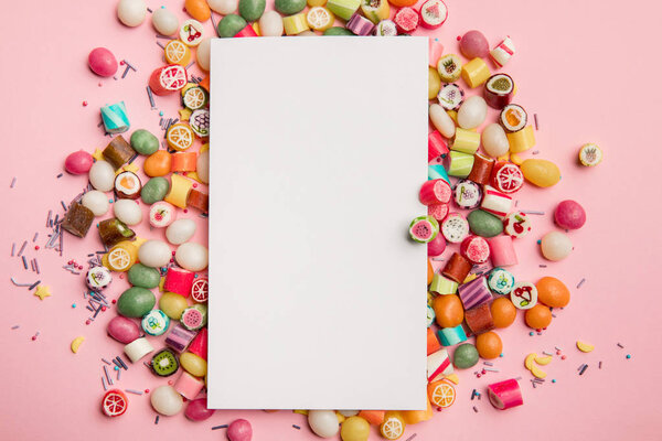 top view of delicious multicolored candies and white blank card with copy space on pink background