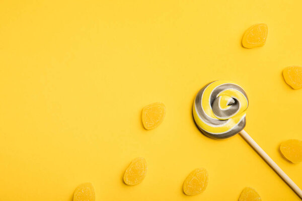 top view of delicious multicolored swirl lollipop on wooden stick and jellies on yellow background