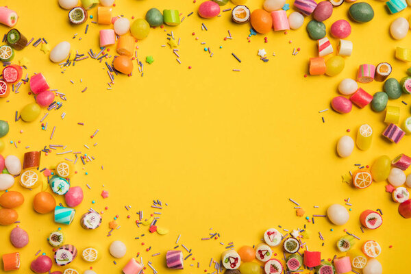 frame of multicolored candies and sprinkles scattered on yellow background with copy space