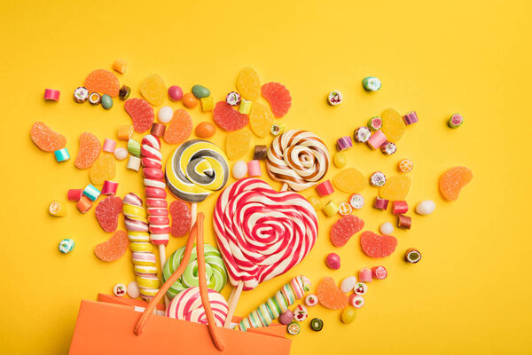 top view of delicious multicolored candies scattered from paper bag on bright yellow background