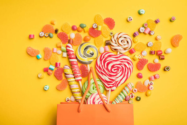 вид сверху на вкусные разноцветные конфеты, разбросанные из бумажного пакета на ярко-желтом фоне

