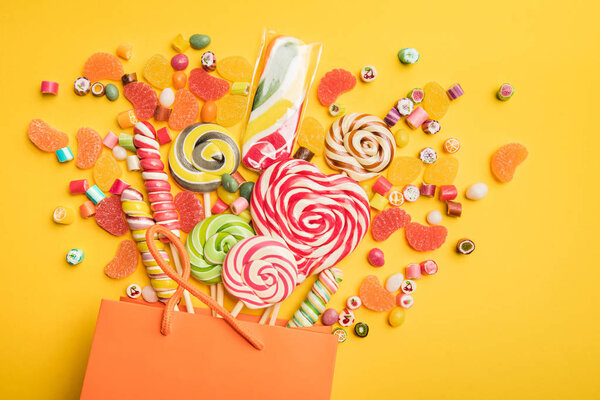 top view of tasty multicolored sweets scattered from paper bag on bright yellow background