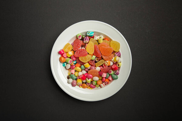 top view of delicious multicolored caramel candies and citrus jellies in white plate on black background 