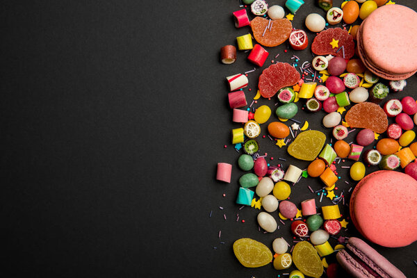 top view of delicious multicolored sweets, sprinkles and macarons on black background 