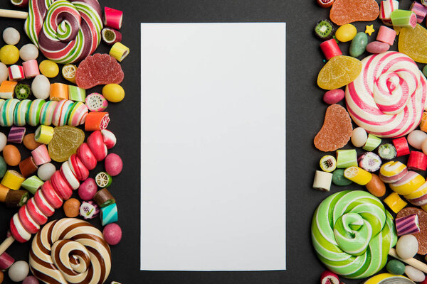 top view of tasty multicolored caramel sweets and lollipops around white empty card on black background with copy space