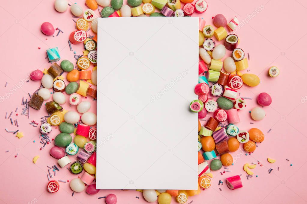 top view of delicious multicolored candies and white blank card with copy space on pink background
