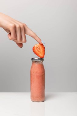 gri üzerinde lezzetli pembe smoothie ile cam şişe yakınında dilimlenmiş çilek tutan kadın kırpılmış görünümü 