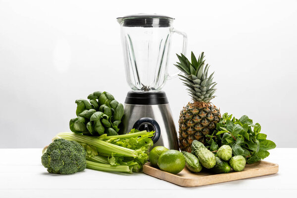 green fresh spinach leaves near organic celery, cucumbers, broccoli fruits and blender on white 