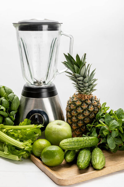 blender near green fresh spinach leaves, organic celery, cucumbers and fruits on white 
