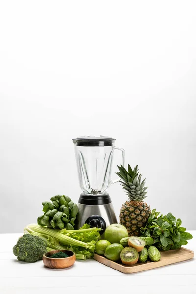 blender near fresh, green vegetables and tasty fruits on white