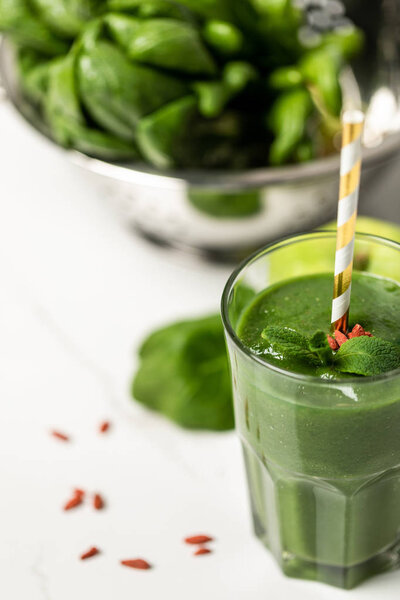 selective focus of tasty smoothie with goji berries and mint in glass with straw near spinach leaves on white 