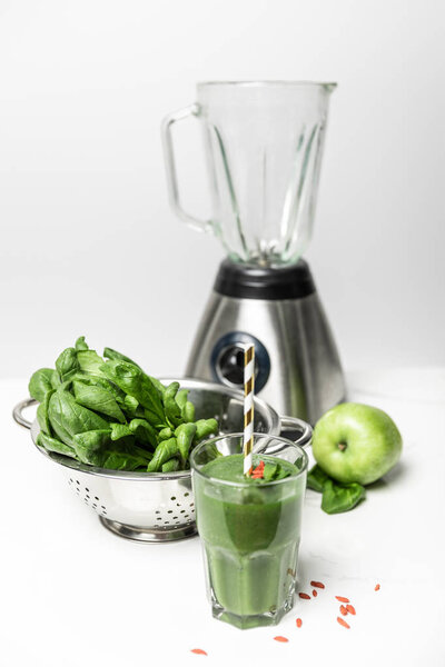 selective focus of  green smoothie in glass with straw near fresh spinach leaves, apple and blender on white 