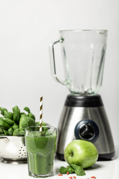selective focus of green smoothie in glass with straw near spinach leaves, apple and blender on white 