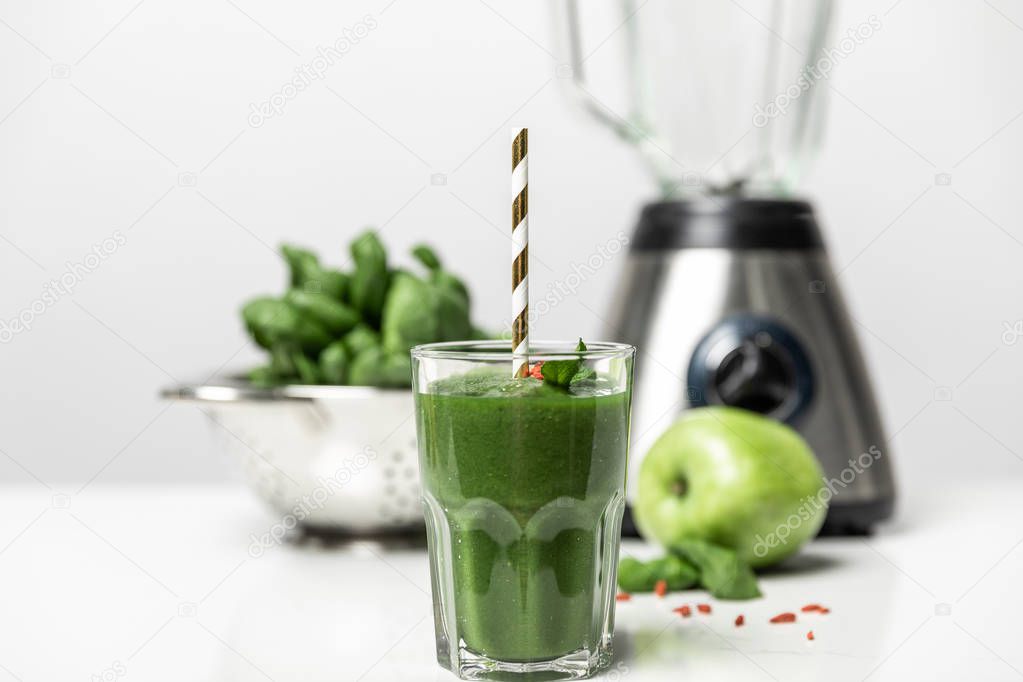 selective focus of tasty smoothie in glass with straw near fresh spinach leaves, apple and blender on white 