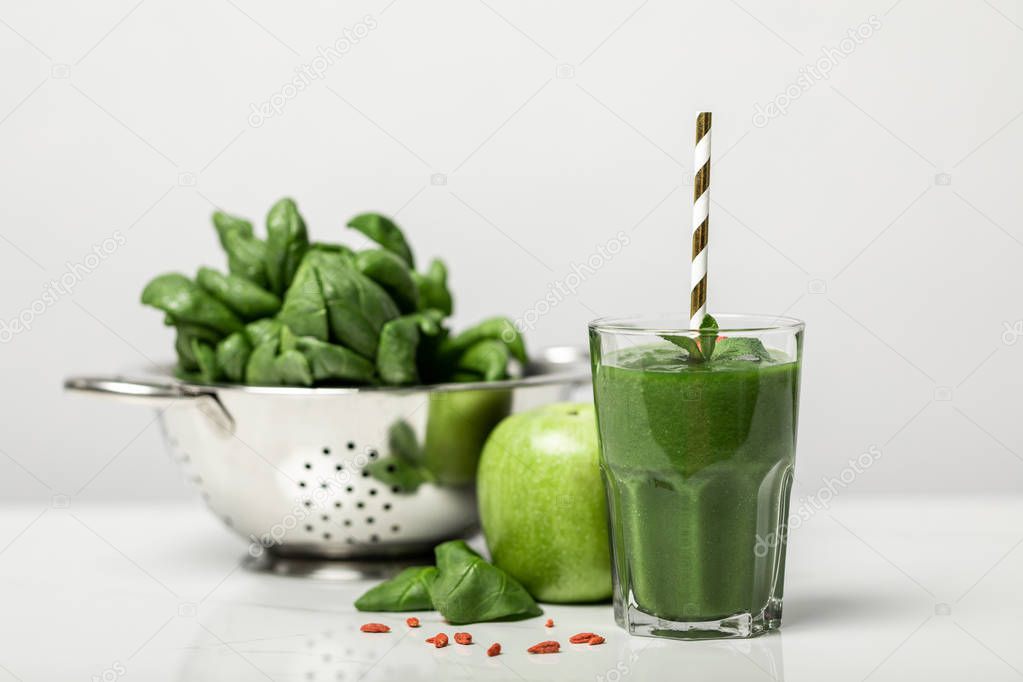 selective focus of green smoothie in glass with straw near spinach leaves and apple on white 