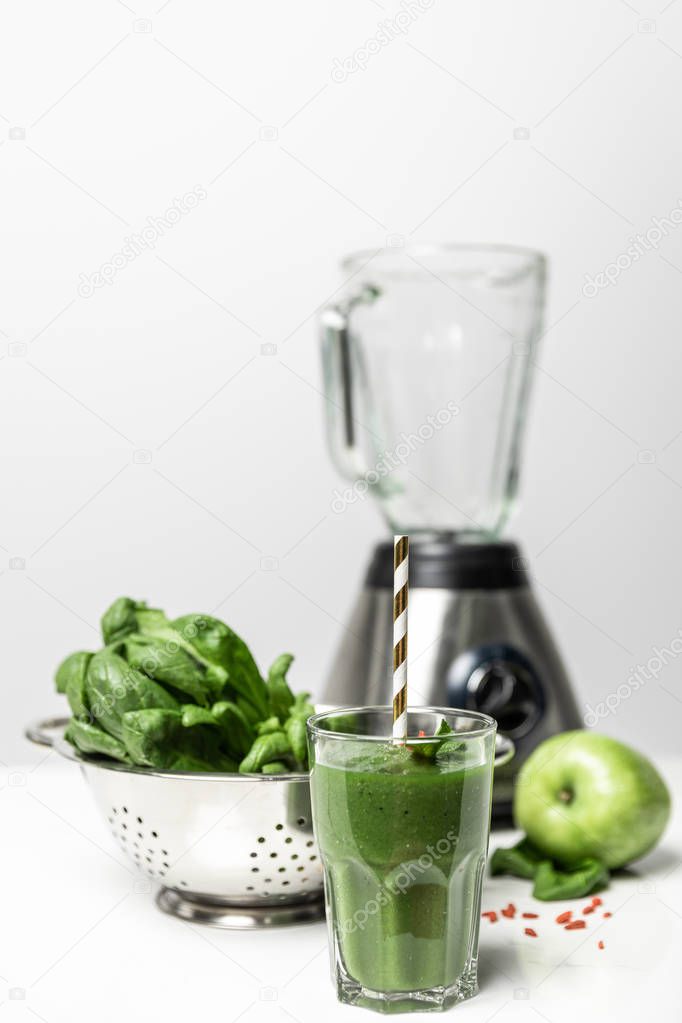 selective focus of green smoothie in glass with straw near fresh spinach leaves, tasty apple and blender on white 