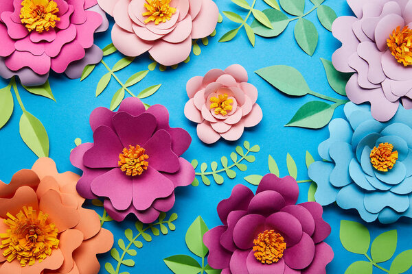 top view of multicolored paper cut flowers with green leaves on blue background