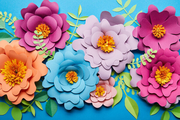 top view of bright colorful paper cut flowers with green leaves on blue background
