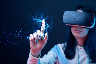 selective focus of young woman in virtual reality headset pointing with finger at glowing cyber illustration on dark background clipart