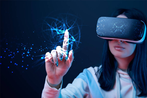 selective focus of young woman in virtual reality headset pointing with finger at glowing cyber illustration on dark background