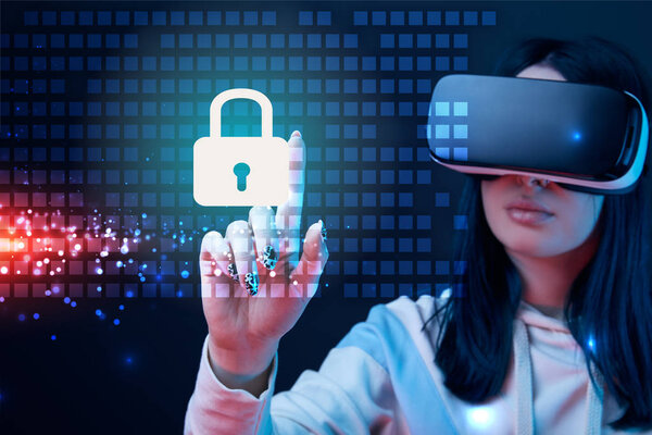 selective focus of young woman in virtual reality headset pointing with finger at glowing cyber security illustration on dark background