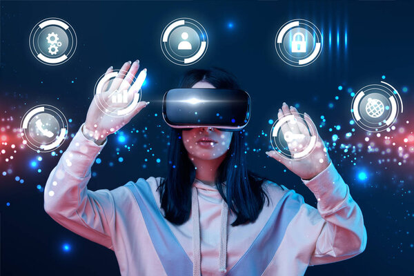 young woman in virtual reality headset gesturing among glowing cyber icons on dark background