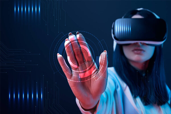 selective focus of young woman in virtual reality headset pointing with hand at cyber illustration on dark background