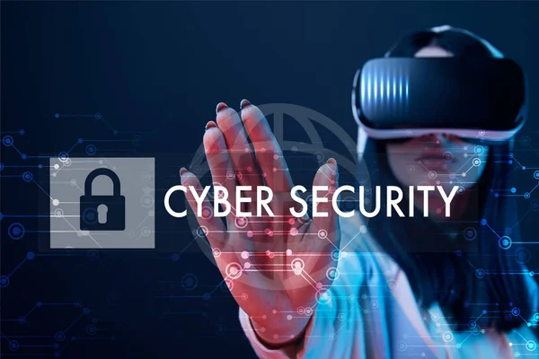 selective focus of young woman in virtual reality headset pointing with hand at cyber security illustration on dark background