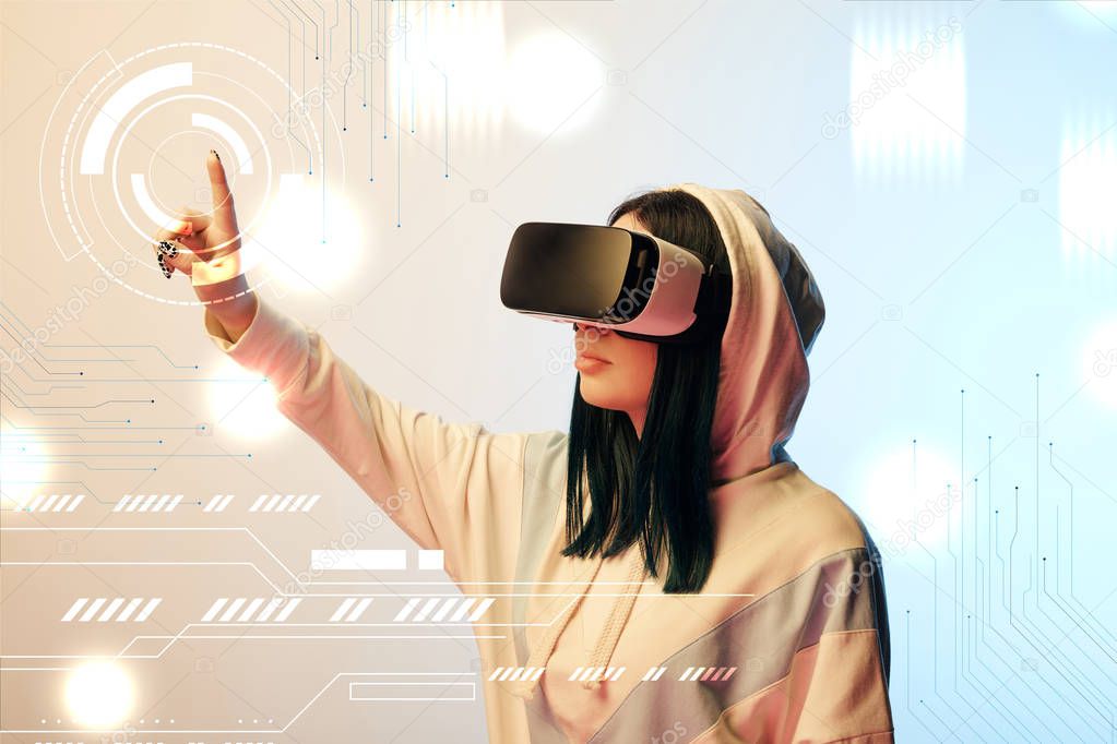 young woman in virtual reality headset pointing with finger at glowing cyber illustration on beige and blue background