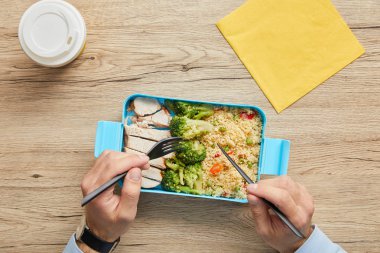 Cropped view of man eating healthy lunch with risotto, broccoli and chicken at wooden table clipart