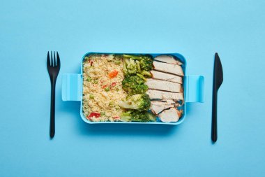 Mavi arka plan üzerinde tek çatal ve bıçak ile risotto, brokoli ve tavuk ile öğle yemeği kutusunun üst görünümü