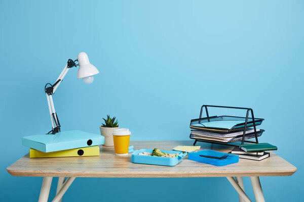 workspace with lunch box and coffee to go on wooden table on blue background