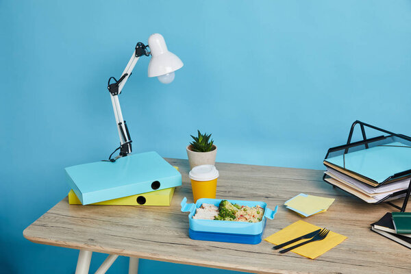 workspace with papers on table and lunch box with healthy food on blue background