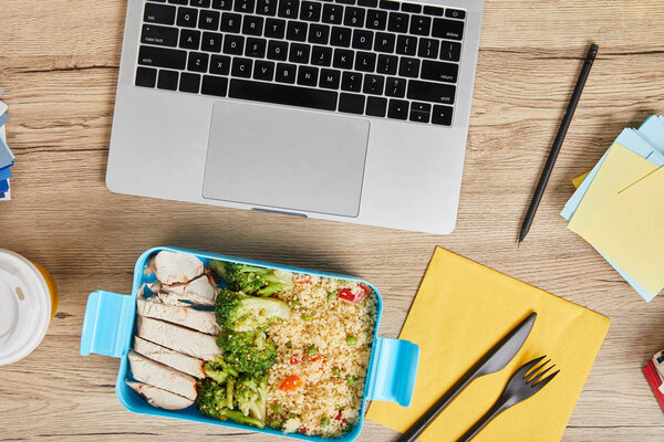 Top view of workplace with laptop and lunch box with tasty risotto and chicken on wooden table, illustrative editorial