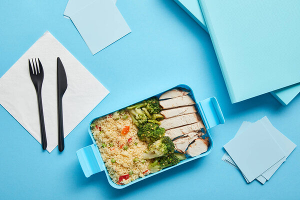 Top view of lunch box with tasty nutritious risotto, chicken and broccoli at workplace on blue background