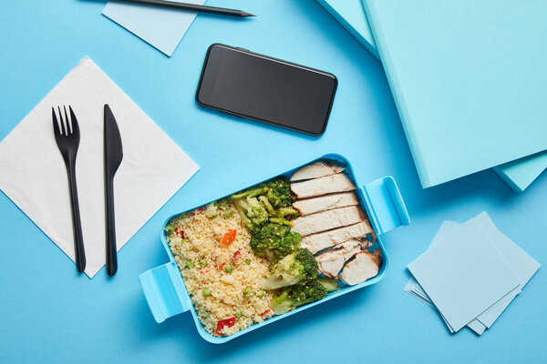 Top view of lunch box with healthy nutritious food and smartphone at workplace on blue background